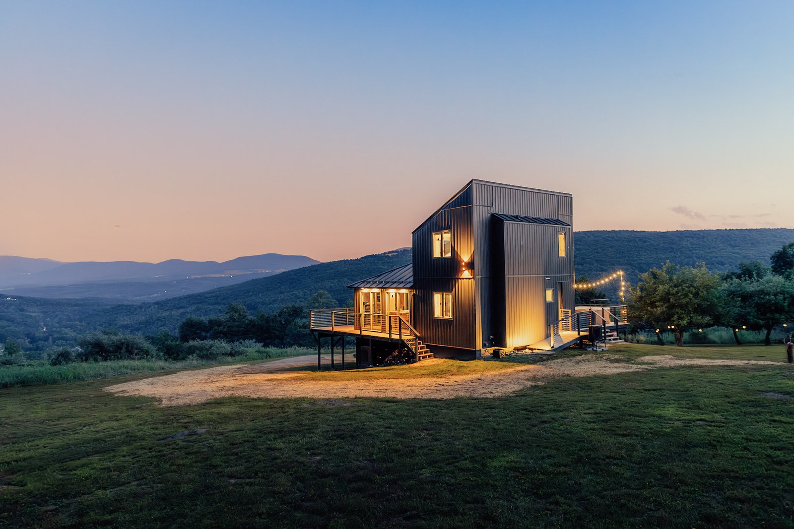 Full exterior establishing shot — house, deck, string lights, Catskill silhouette