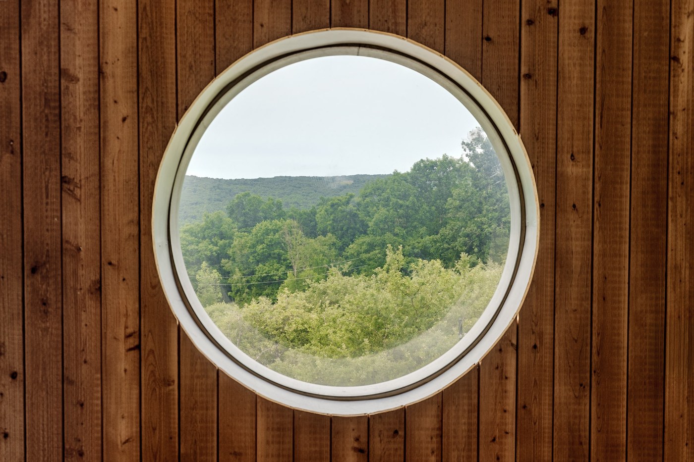 Round porthole window from inside, framed by wood paneling with mountain and treeline view